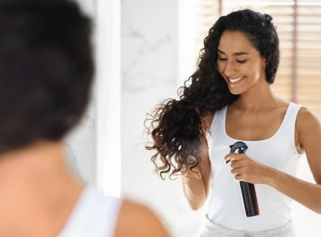 Haircare. Beautiful Young Lady Applying Hair Spray, Standing Near Mirror In Bathroom